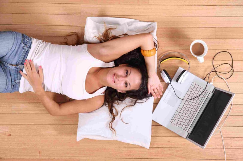 Estudiante recostada en el piso con laptop, audífonos y café, disfrutando un momento de estudio relajado.