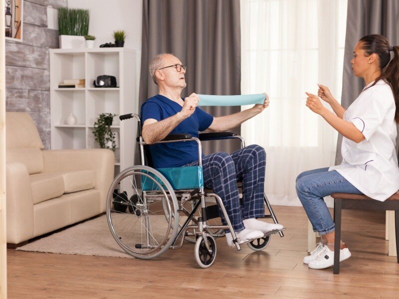 Fisioterapeuta trabajando con un hombre mayor en silla de ruedas usando una banda elástica, fortaleciendo músculos y promoviendo la rehabilitación.