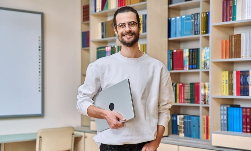 Docente con computadora en biblioteca que busca estudiar una maestría en Educación para continuar su desarrollo profesional.