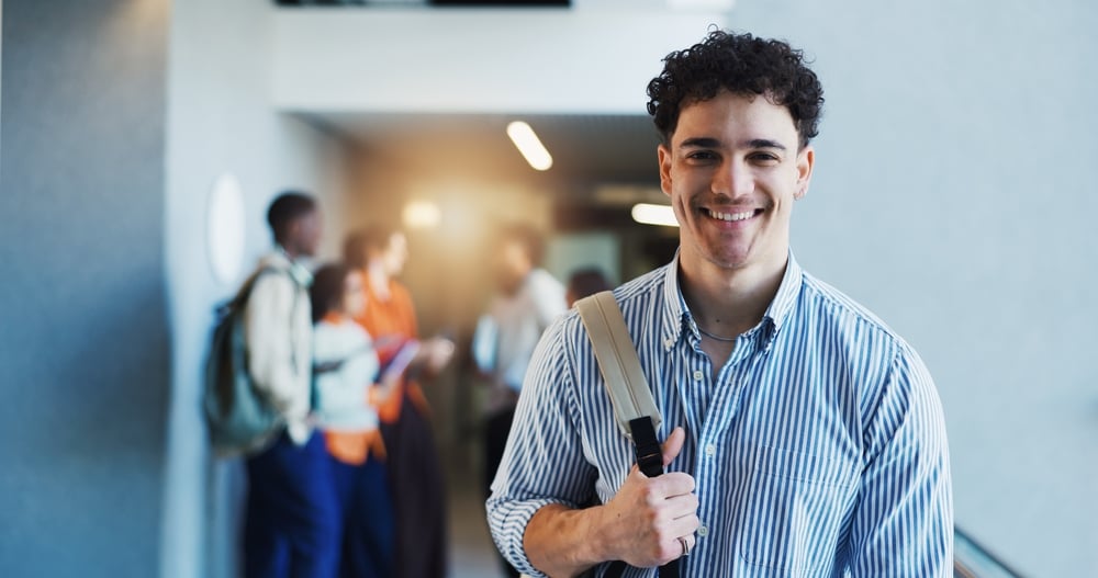 Joven sonriente con mochila en un pasillo, con otros estudiantes al fondo.